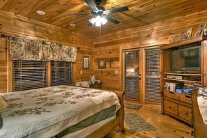 A wooden cabin bedroom with a bed, a ceiling fan, a television, and a bookshelf.