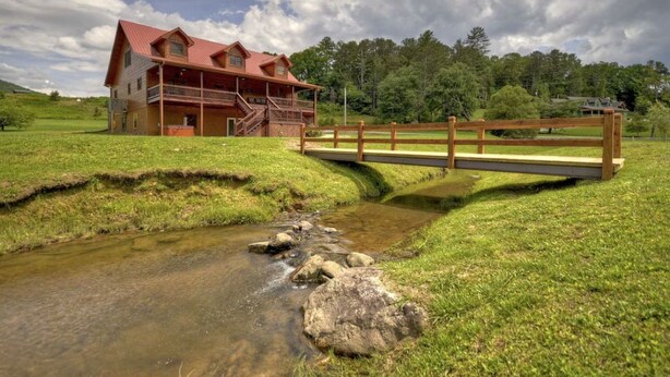 A wooden house with a red roof, a wooden bridge over a stream, and a grassy landscape.