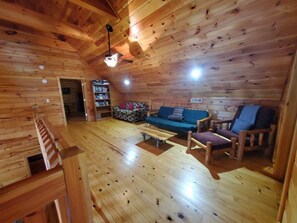 L’intérieur d’un chalet en bois comprend une chambre à coucher située en mezzanine, un espace de vie avec un canapé et des fauteuils, ainsi qu’une bibliothèque.