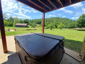 Un bain à remous sous une terrasse couverte, avec vue sur un paysage verdoyant et un chalet en bois au loin.