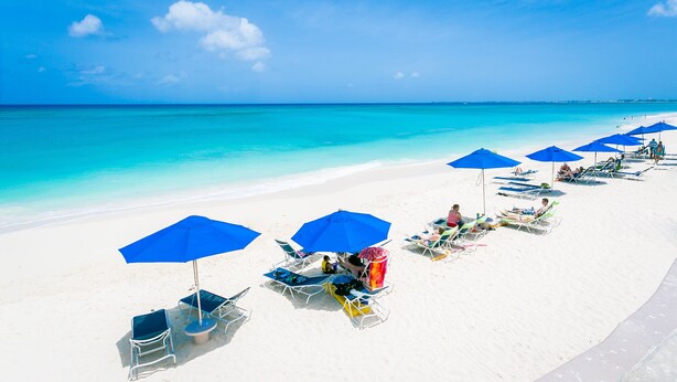 Una playa con arena blanca y agua turquesa cristalina. Se observan sombrillas azules y sillones de playa dispuestos a lo largo de la orilla.