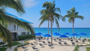 Un resort frente a la playa con una fila de tumbonas bajo sombrillas azules, palmeras y vista al océano.