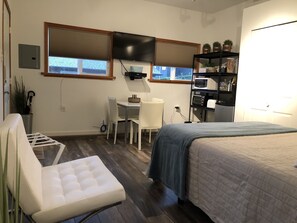 A compact bedroom with a bed, a desk, and a chair. There is a TV mounted on the wall, a shelf with kitchen appliances, and a window with blinds. - One Bedroom in West Vancouver (West Vancouver)