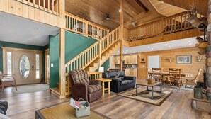 A spacious living area with a high ceiling, wooden staircase, and a view of the kitchen.
