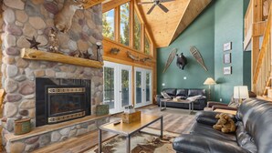 A cozy living room with a stone fireplace, wooden beams, a leather couch, and a large window overlooking the outdoors.