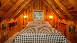Une chambre mansardée chaleureuse, avec une couvre-lit à carreaux, des murs en bois, une fenêtre et deux lampes fixées au mur.
