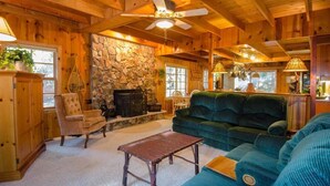 L’intérieur chaleureux d’un chalet avec une cheminée en pierre, des murs en bois, un canapé vert et un ventilateur de plafond.