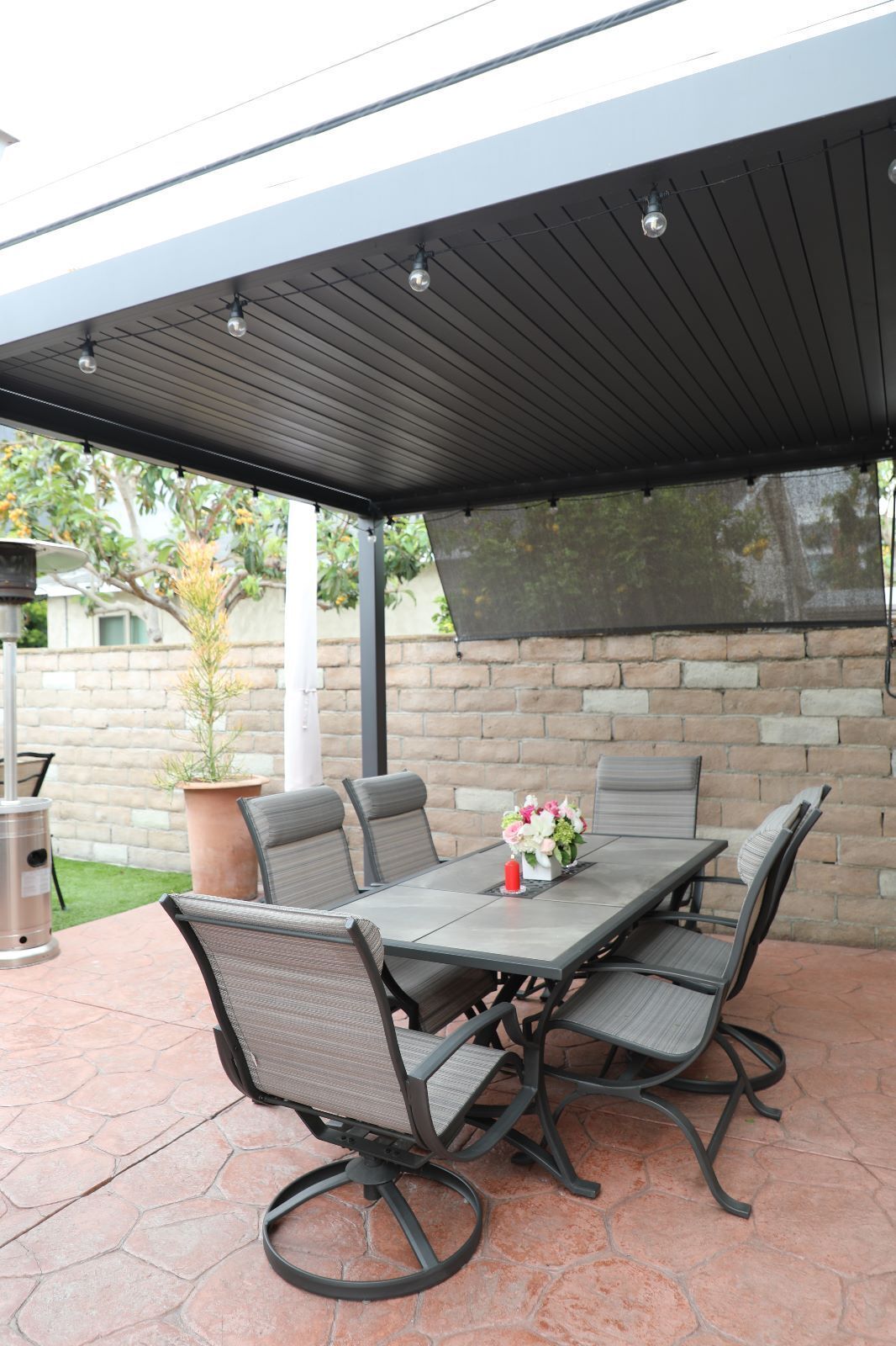 An outdoor patio with a covered table and chairs, a potted plant, and a brick wall in the background.