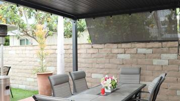 An outdoor patio with a covered table and chairs, a potted plant, and a brick wall in the background.