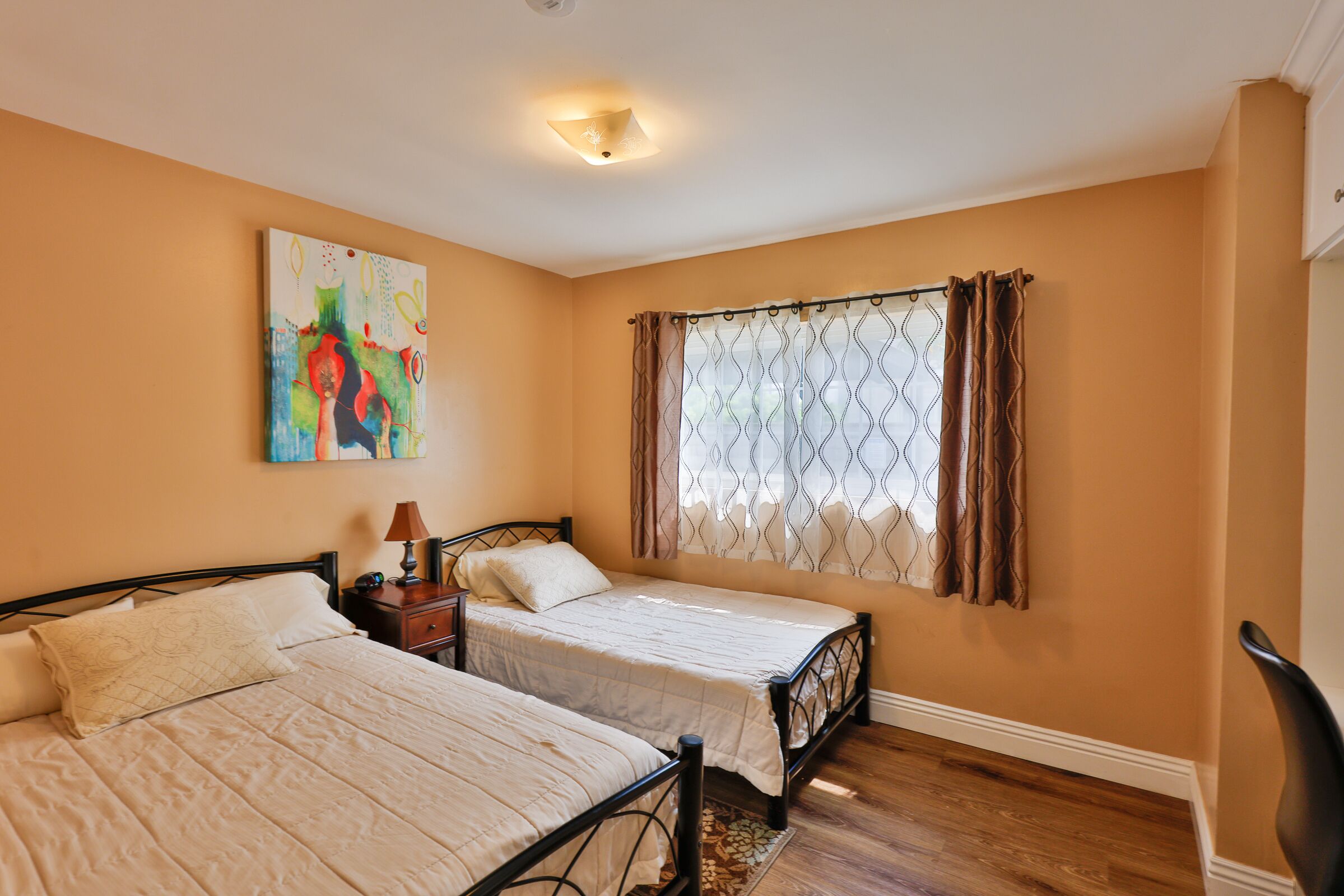 A bedroom with two beds, a nightstand, a lamp, a painting, and a window with curtains.