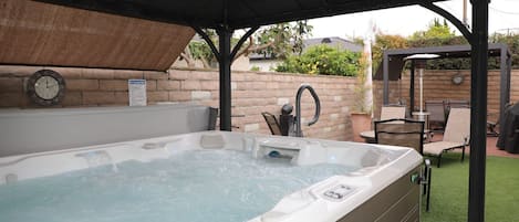 A hot tub under a covered patio with a brick wall, outdoor furniture, and a clock on the wall.