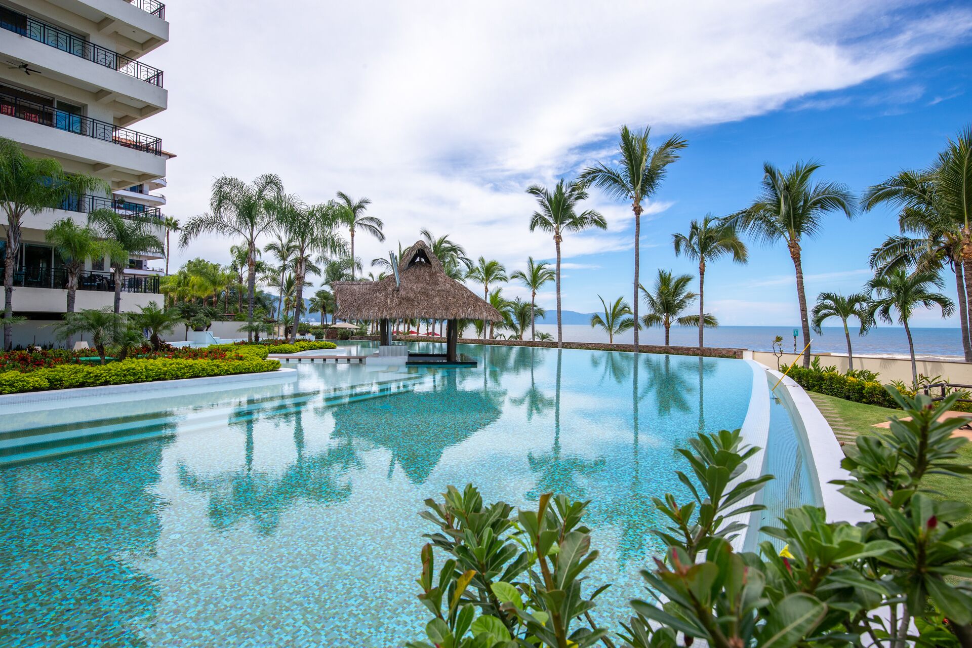Een zwembad van een resort met een rieten hut, palmbomen en uitzicht op de oceaan.
