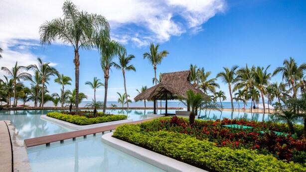 Zwembadgedeelte van een resort met helderblauw water, omgeven door palmbomen en een constructie met rieten dak.