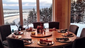 Une table de salle à manger ronde, entourée de chaises, avec des assiettes remplies de nourriture et une cafetière, dans une pièce dotée de grandes fenêtres donnant sur un paysage enneigé.