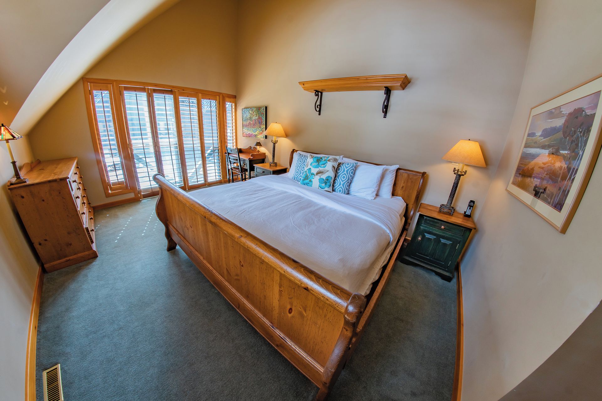 A bedroom with a large bed, wooden headboard, two bedside tables with lamps, a wooden dresser, and a window with blinds.