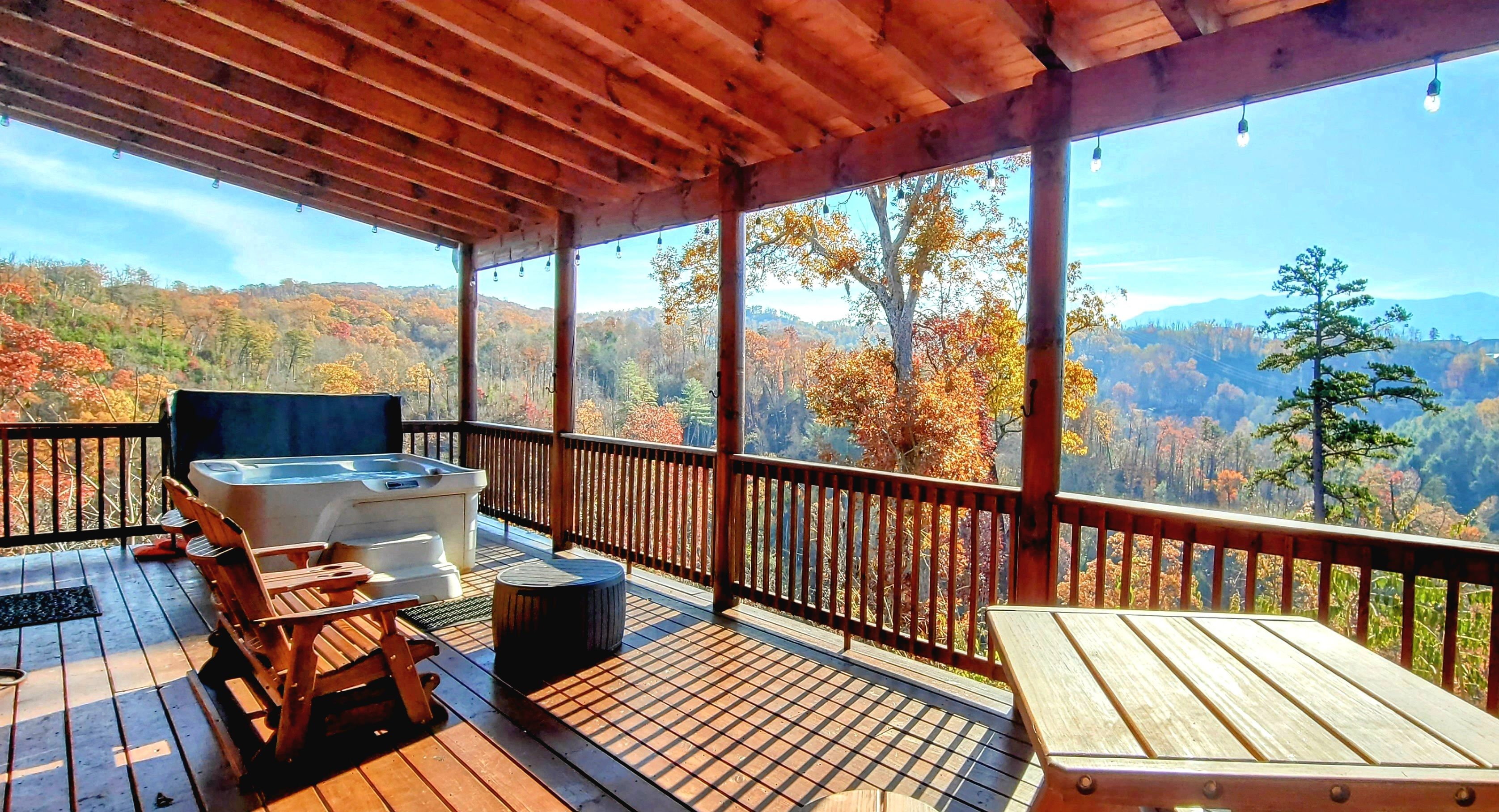 A wooden deck with a hot tub, chairs, and a table overlooking a forested valley.