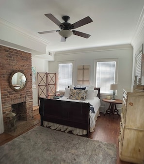 A bedroom with a bed, a ceiling fan, a brick wall, a fireplace, and a window with blinds.