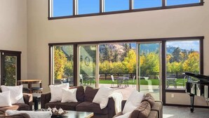 A spacious living room with a sectional sofa, a piano, and large windows offering a view of a landscaped garden. - The Leavenworth Lodge - Swimming Pool, Pickleball (Leavenworth)