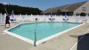 Une piscine rectangulaire à l’eau d’un bleu limpide, entourée de chaises et de parasols blancs. Une personne nettoie le bord de la piscine.