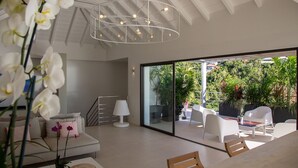 A modern living room with a sofa, a coffee table, and a chandelier. There is a sliding glass door leading to an outdoor patio with chairs and plants. - NEW Villa AKA walking distance to the beach (Saint Barthelemy)