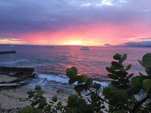 Un coucher de soleil sur une mer calme, avec des bateaux, une jetée et une végétation au premier plan.