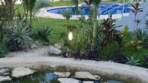 En swimmingpool med oregelbundet formade stenar, omgiven av olika tropiska växter och en vit byggnad i bakgrunden.