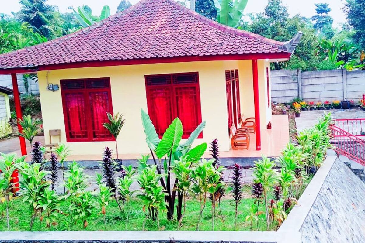 A single-story house with a red-tiled roof, a porch with a red railing, and a small garden with various plants.