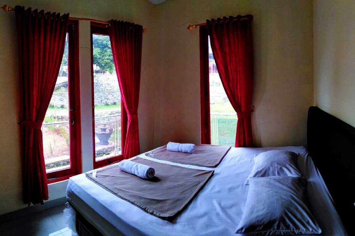 A double bed with two pillows and a rolled towel, red curtains, and a view of greenery outside.