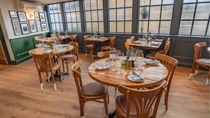 Un restaurante con mesas y sillas de madera redondas, ventanas amplias con persianas e imágenes enmarcadas en la pared.