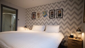 Una cama bien tendida con sábanas blancas, una mesita de noche de madera con una lámpara, y una pared con diseño en zigzag.