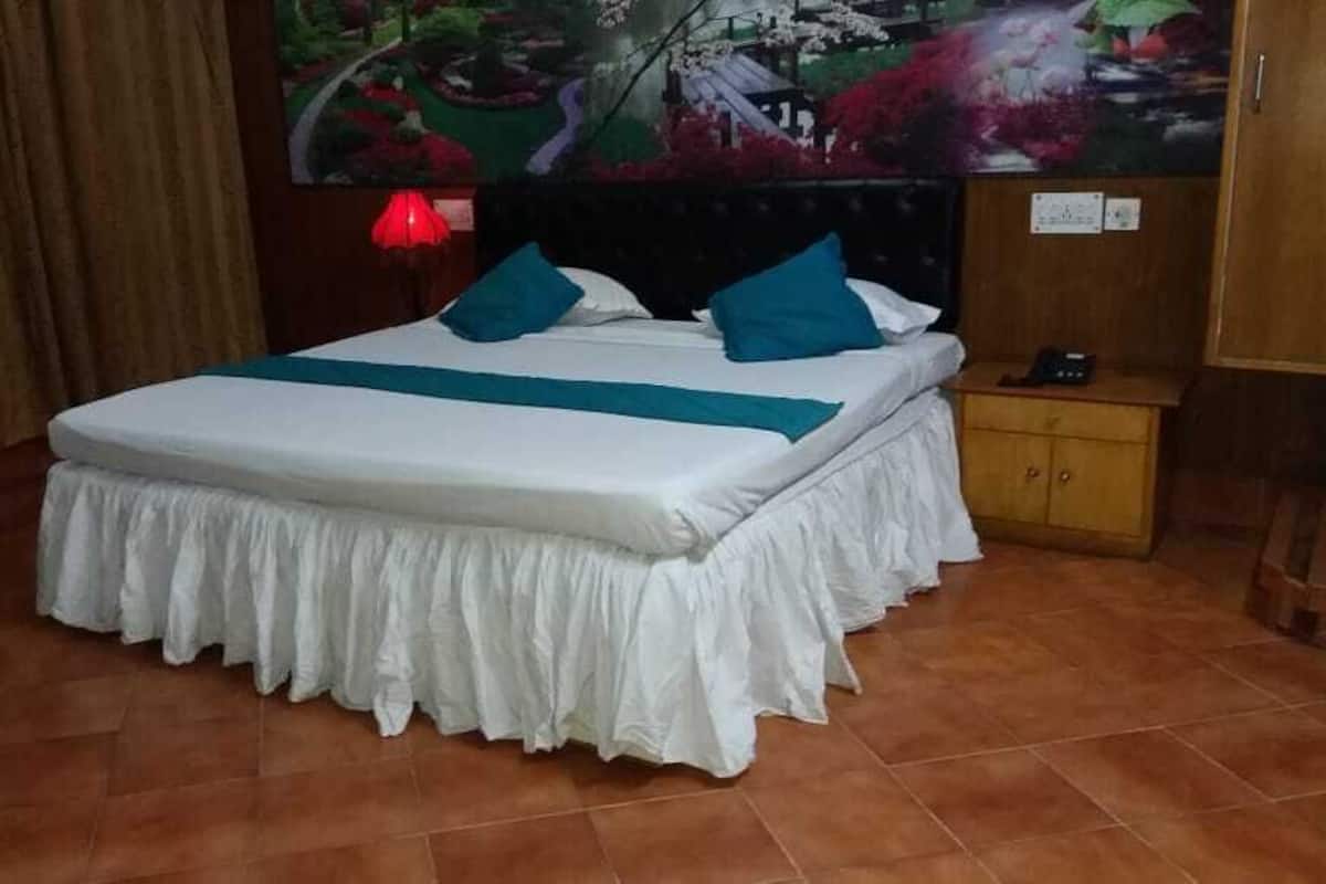 Un lit bien fait, recouvert d’une couvre-lit blanche agrémentée de touches turquoise, une table de chevet en bois avec une lampe, et un grand tableau floral accroché au mur.