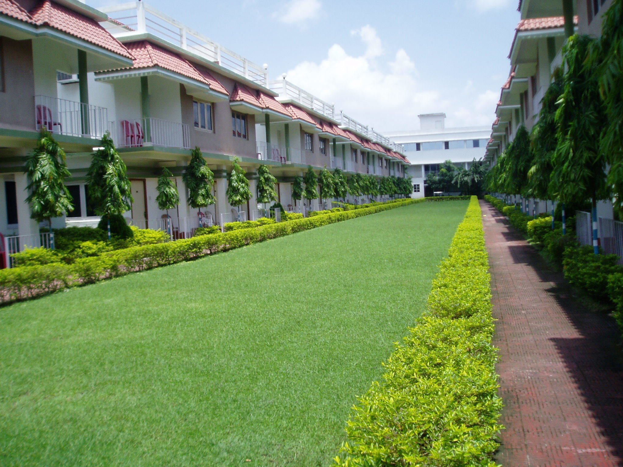 A hotel with a long, straight pathway flanked by neatly trimmed hedges and a row of balconies with red-tiled roofs.