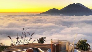 A table with a tray of food and two glasses of a dark beverage, set against a backdrop of a mountain and clouds. - N'jung Bali Camp (Kintamani)