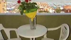 Un balcon avec une table et des chaises, donnant sur une plage parsemée de parasols et sur une jetée.
