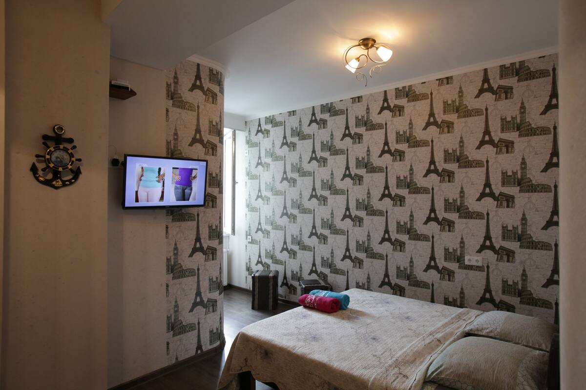 A hotel room with a bed, a television on the wall, and a patterned wallpaper featuring the Eiffel Tower.