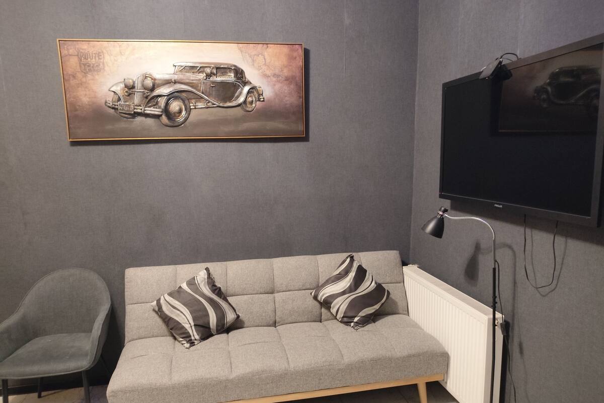 A modern living room with a grey sofa, two striped pillows, a grey armchair, a mounted television, and a framed artwork of a vintage car.