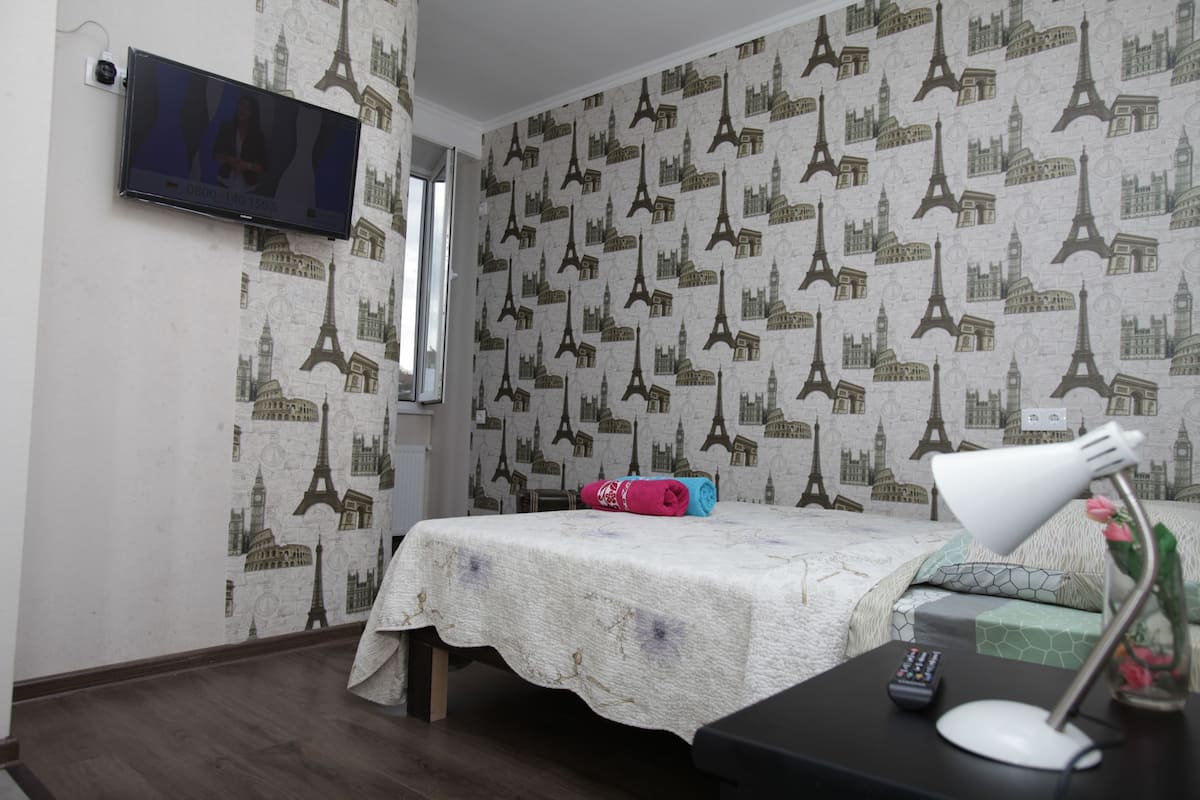 A room with a bed, a desk with a lamp, a TV mounted on the wall, and a patterned wallpaper featuring the Eiffel Tower.