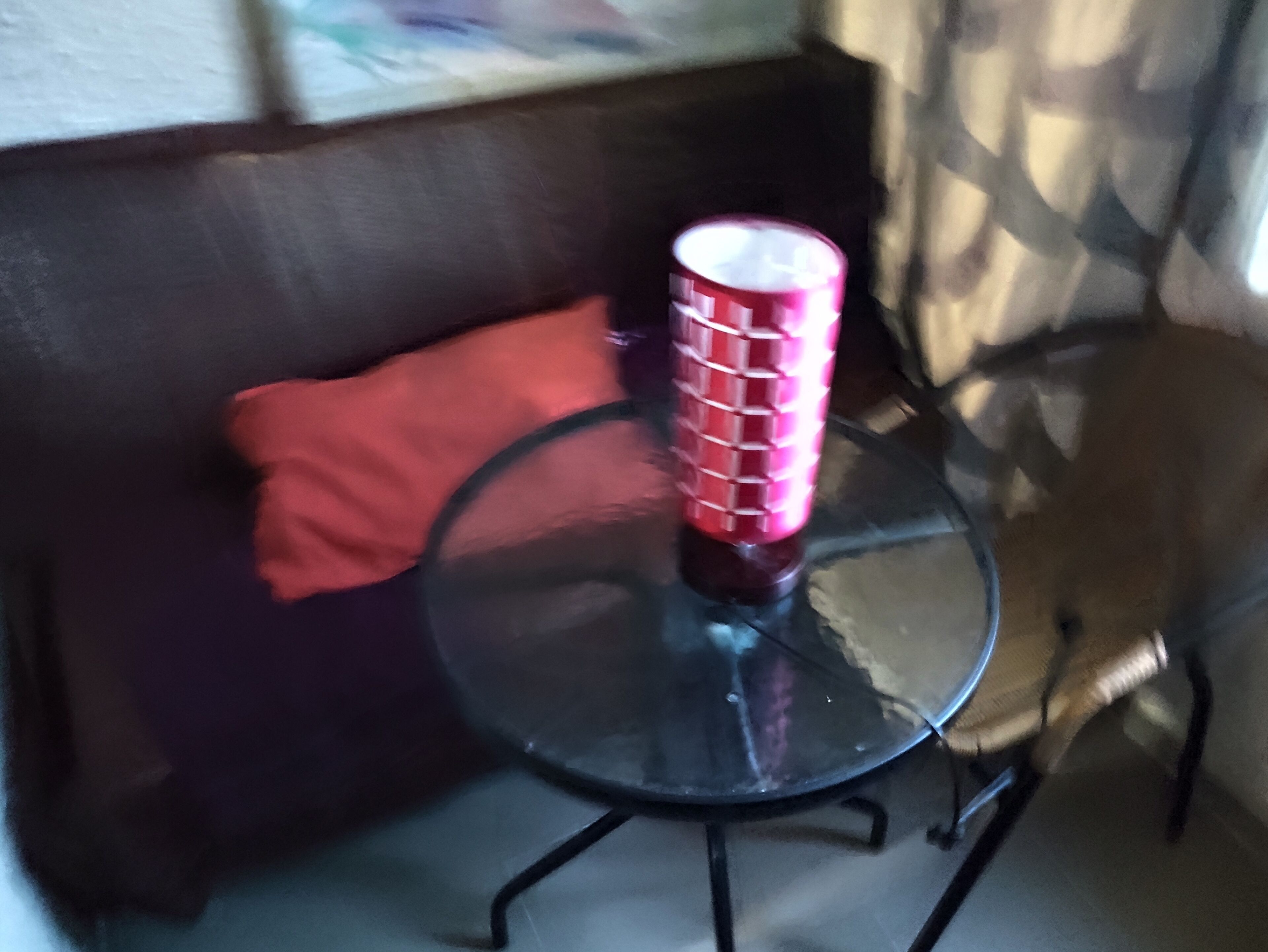 Une table en verre sur laquelle repose un objet cylindrique rouge, un coussin rouge sur un canapé sombre, et un mur à motifs en arrière-plan.