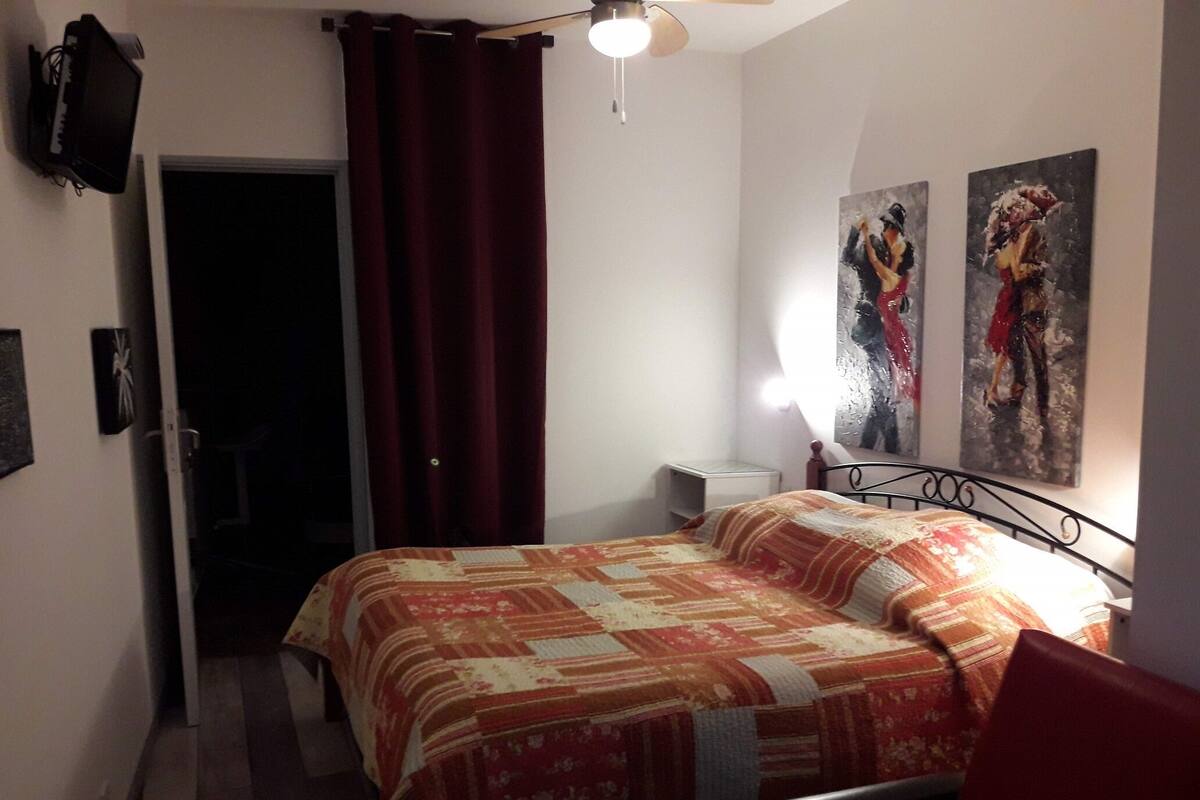 A bedroom with a bed, a nightstand, a lamp, a ceiling fan, and two paintings on the wall.