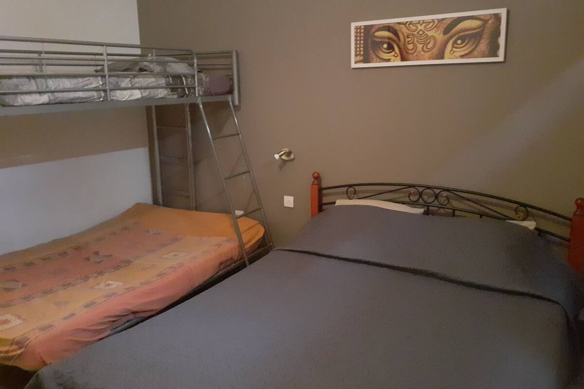 A dormitory room with two beds, one with a bunk bed and the other with a single bed. There is a ladder against the wall, a painting on the wall, and a door with a handle.
