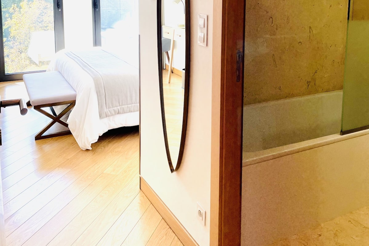 A modern bedroom with a large bed, a bedside table, and a large oval mirror on the wall. There is a glass shower enclosure to the right.