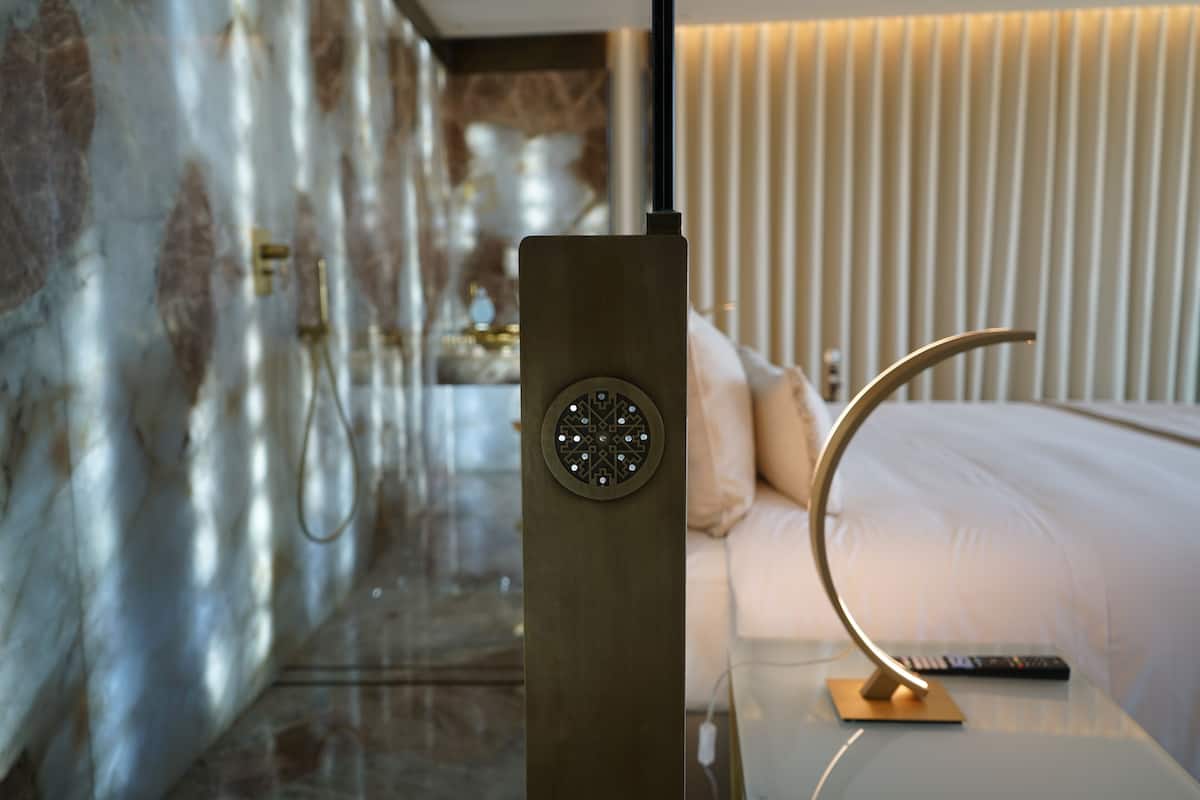 A modern hotel room with a marble wall, a bed with white linens, a bedside table with a remote control, and a unique wall-mounted light fixture.