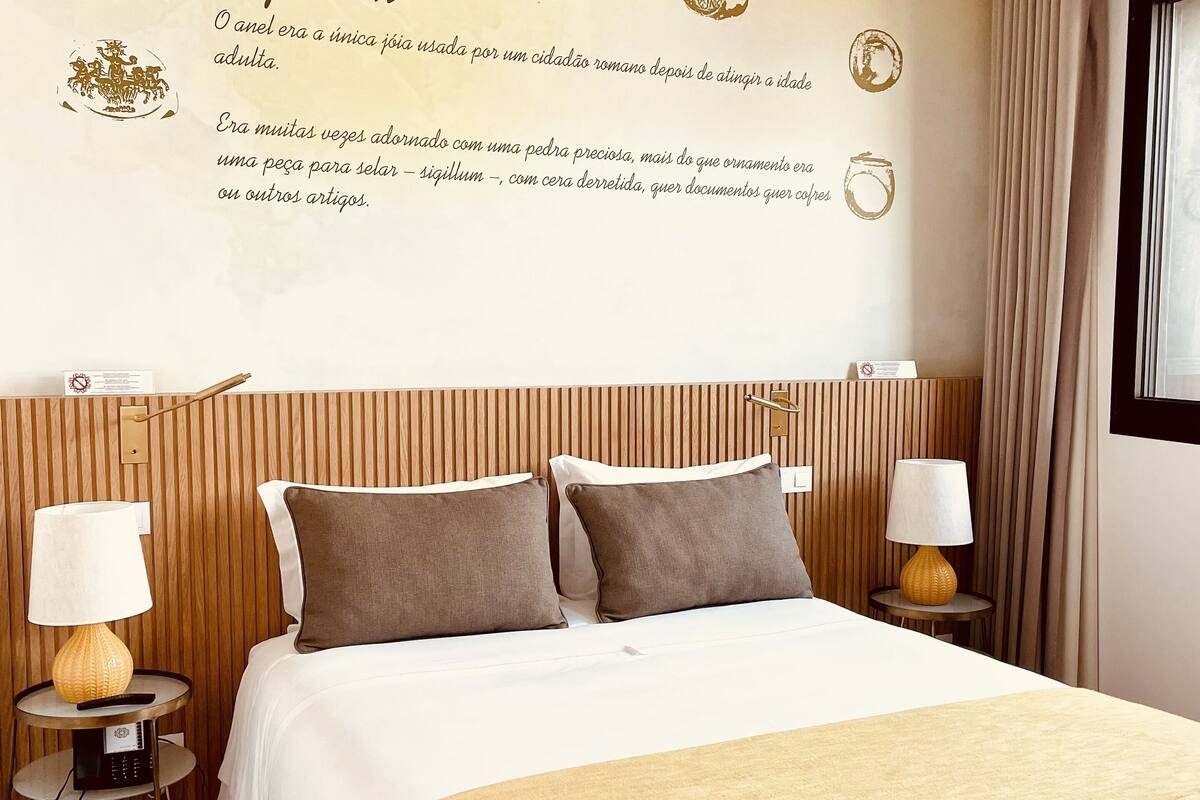 A hotel room with a large bed, two bedside tables with lamps, a wall with decorative text and images, and a window with curtains.