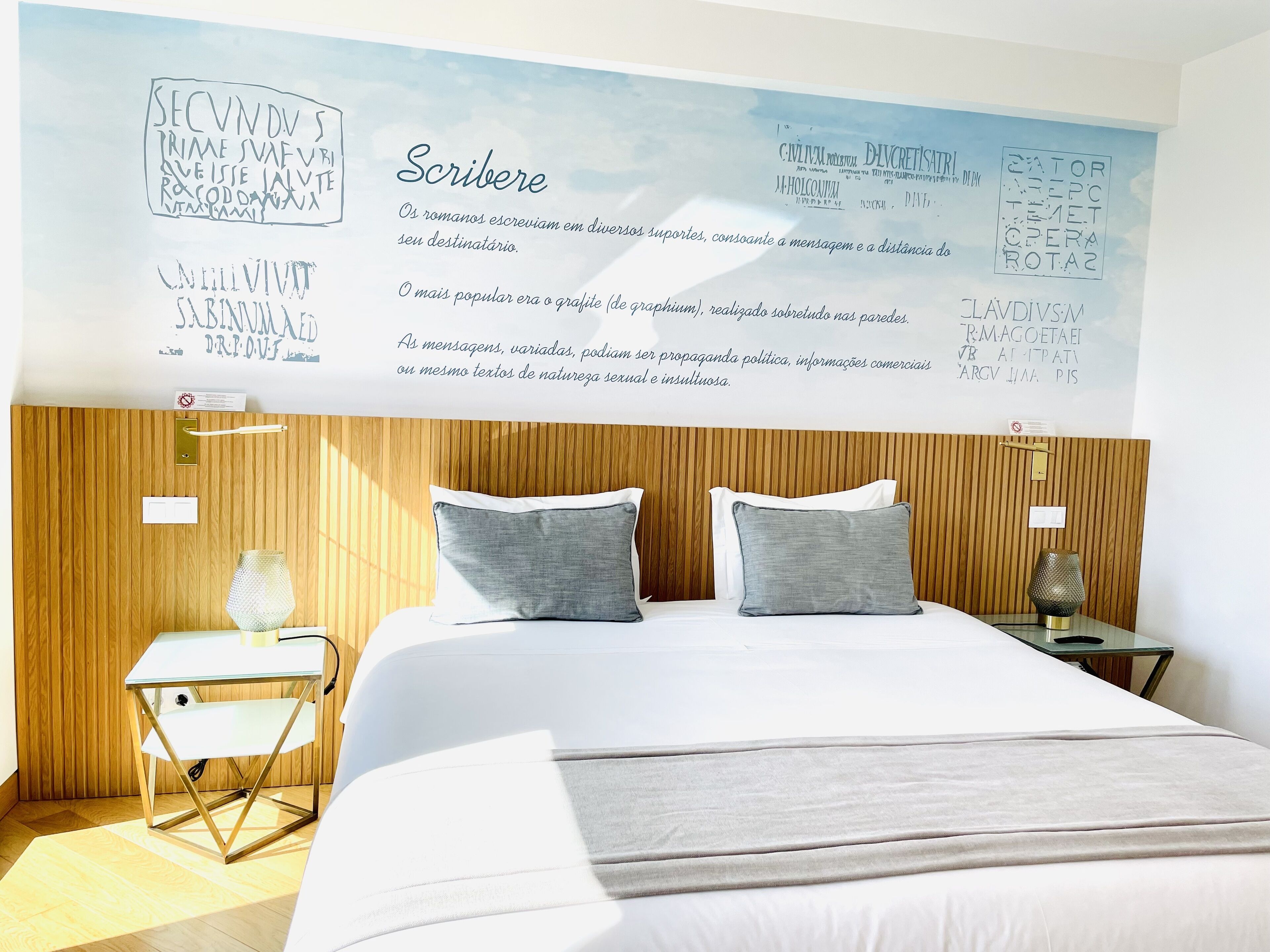 A modern hotel room with a large wall mural, a wooden headboard, a bed with grey bedding, and a small bedside table with a lamp.