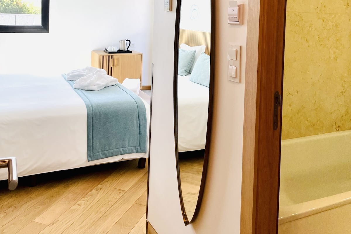 A hotel room with a bed, a bedside table, a mirror, and a view of the outside.