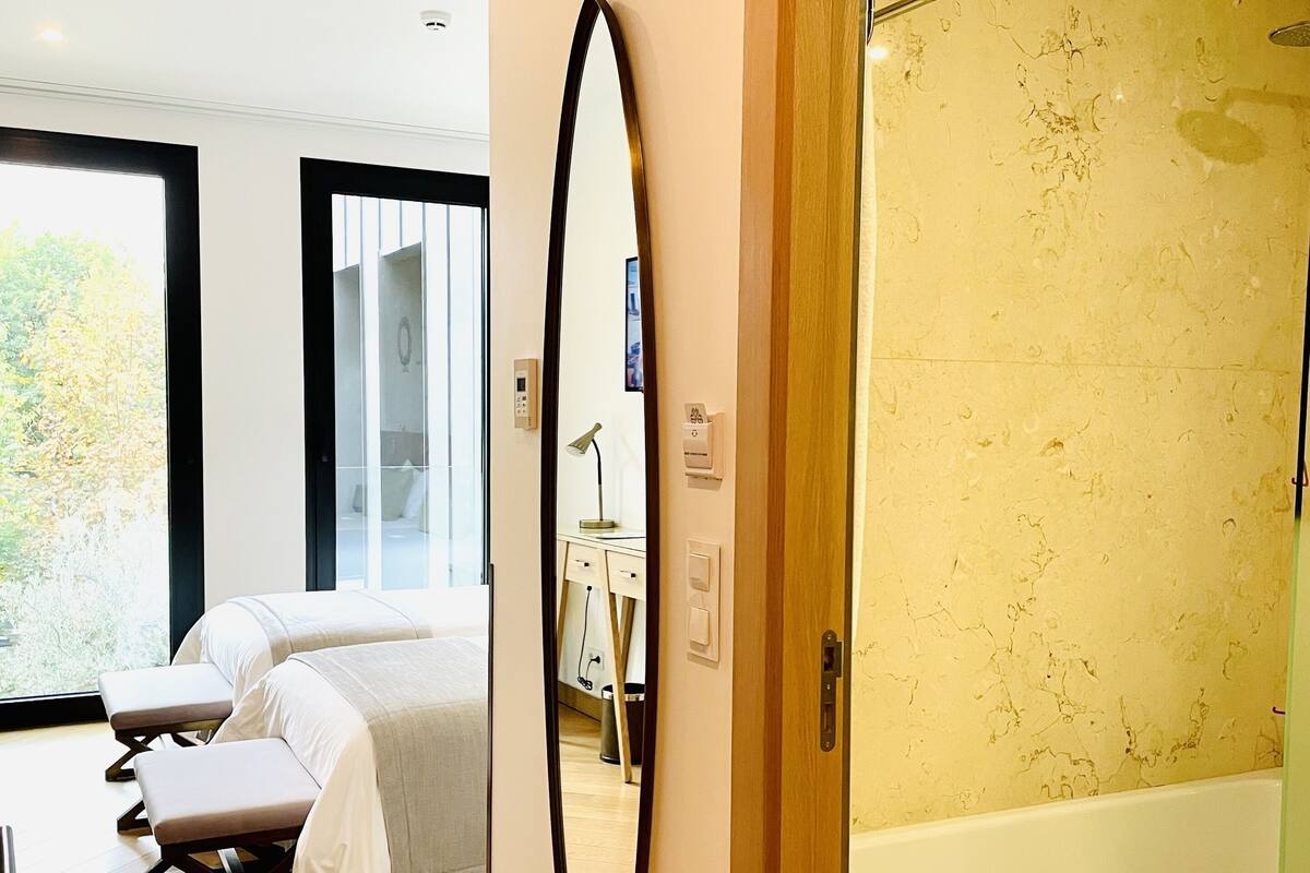 A modern bedroom with a large bed, a bench, a mirror, and a bathroom with a bathtub and shower.