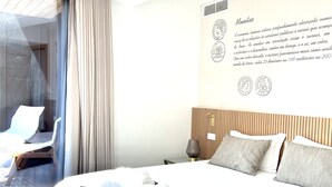 A modern hotel room with a large bed, a sitting area with a chair and a table, a view through a glass door, and a wall with informational text and coin images. - Castelo Hotel (Chaves)