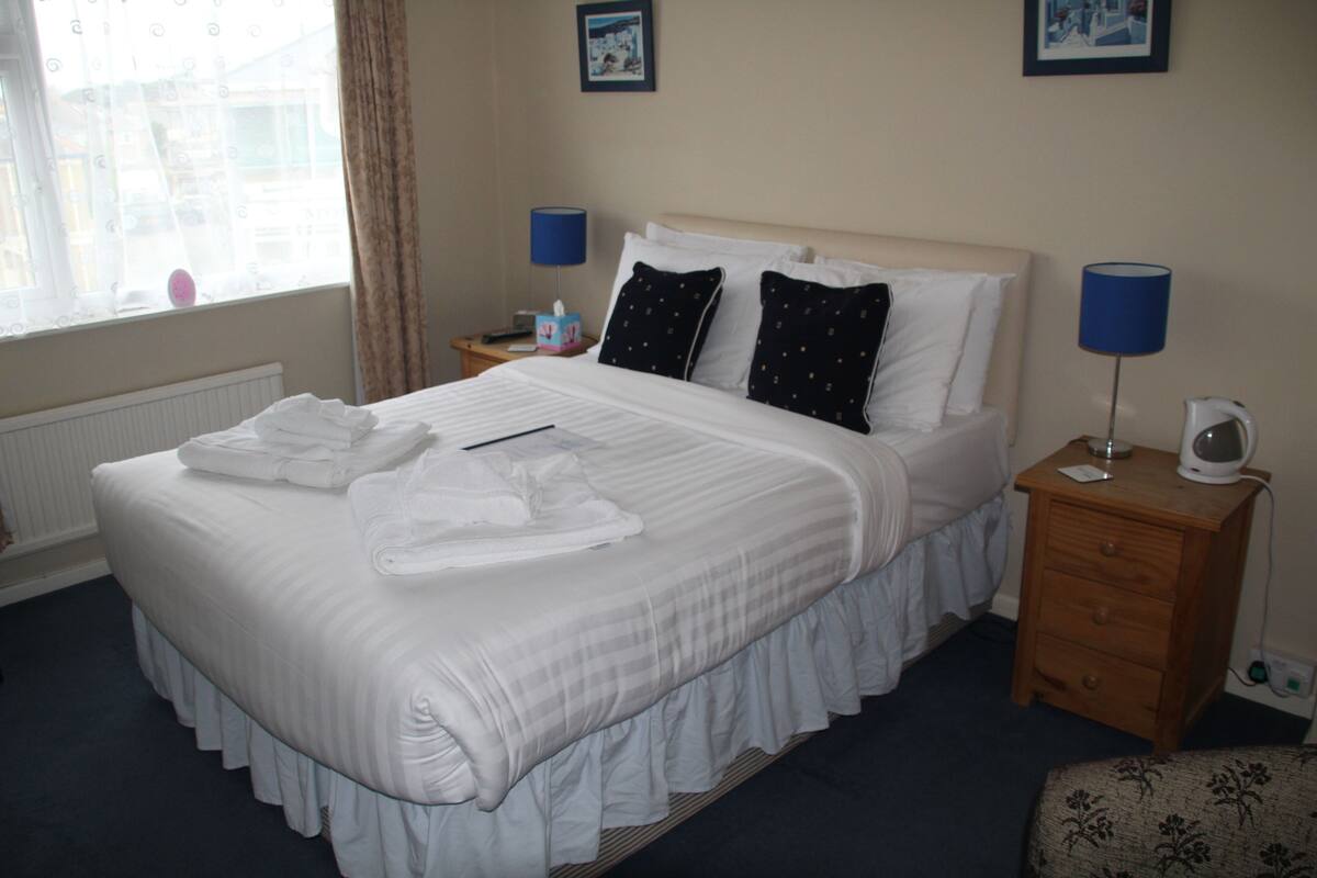 A neatly made bed with white linens and towels, a bedside table with a lamp, and a small drawer.