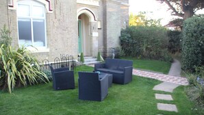 A well-maintained garden with a paved walkway, outdoor furniture, and a brick building with a green door. - The Kenbury (Shanklin)