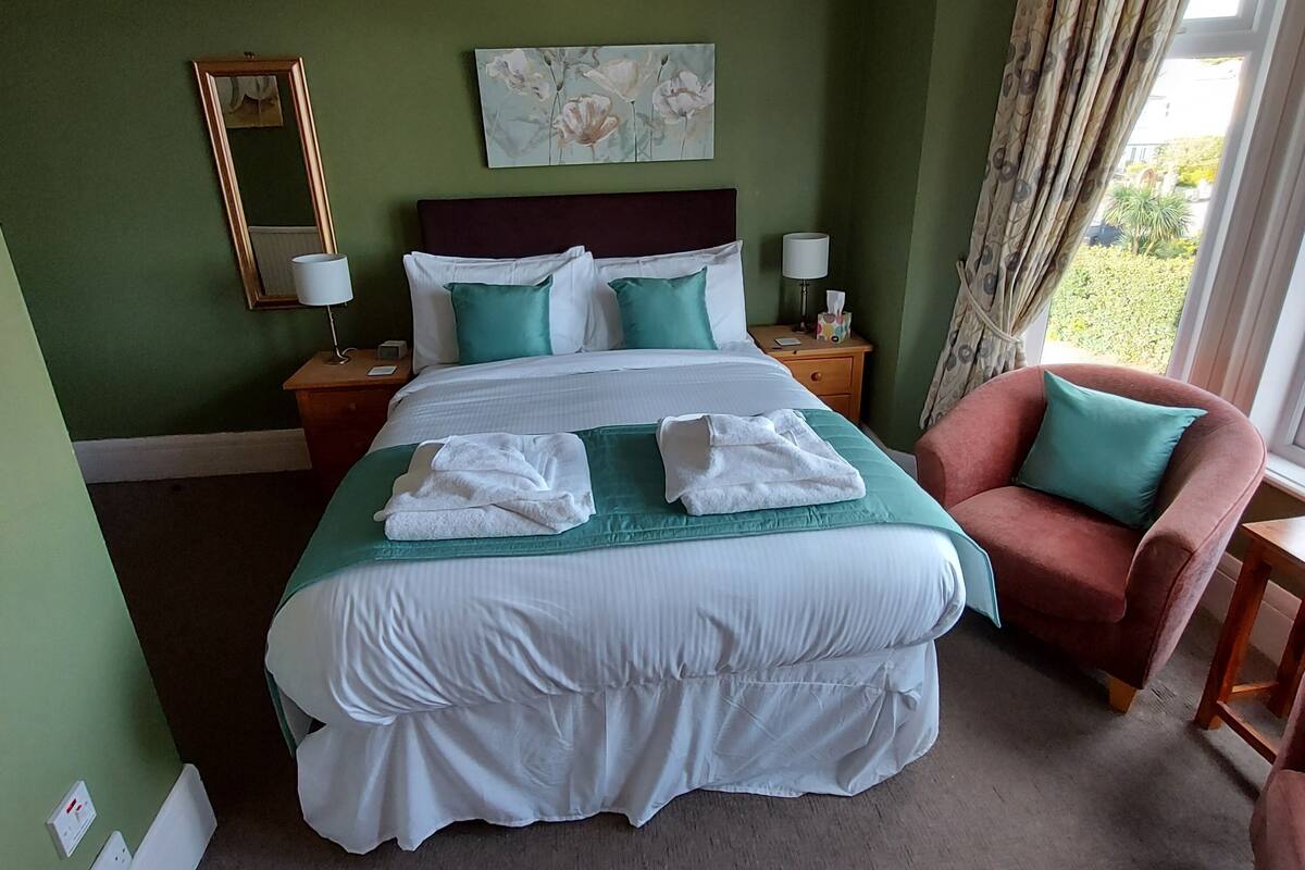 A neatly made bed with white linens, a green accent pillow, and two folded towels on a green bedspread. There is a wooden nightstand with a lamp and a tissue box beside the bed. A red armchair is positioned to the right, and a window with curtains is visible in the background.